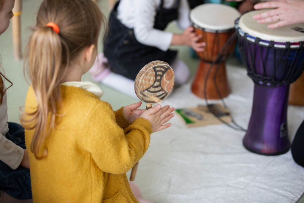 A little girl playing maracas, with others playing hand drums during an indoor music activity.